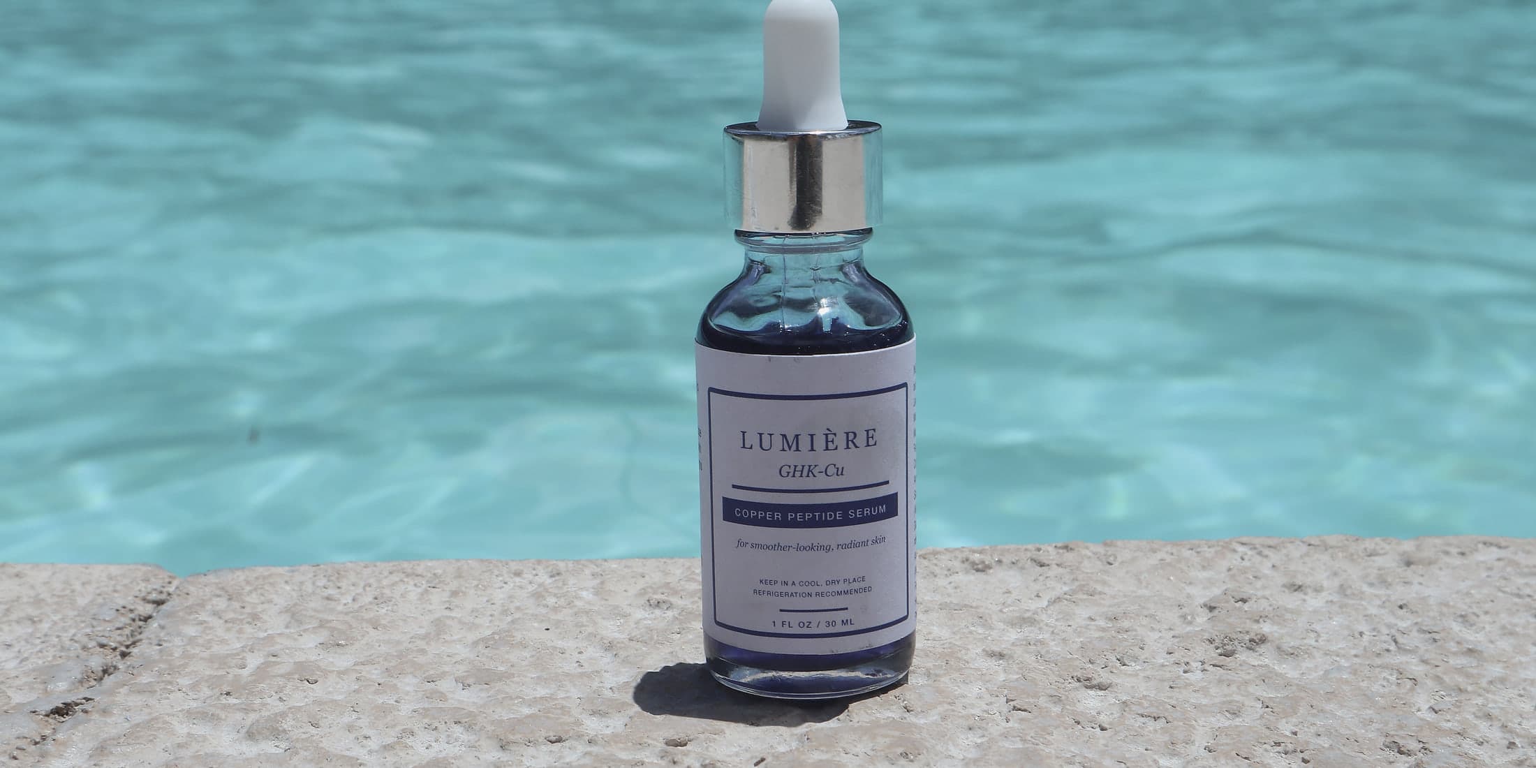 Lumiere Routine GHK-Cu Copper Peptide Serum standing on sunlit travertine at the edge of a pool