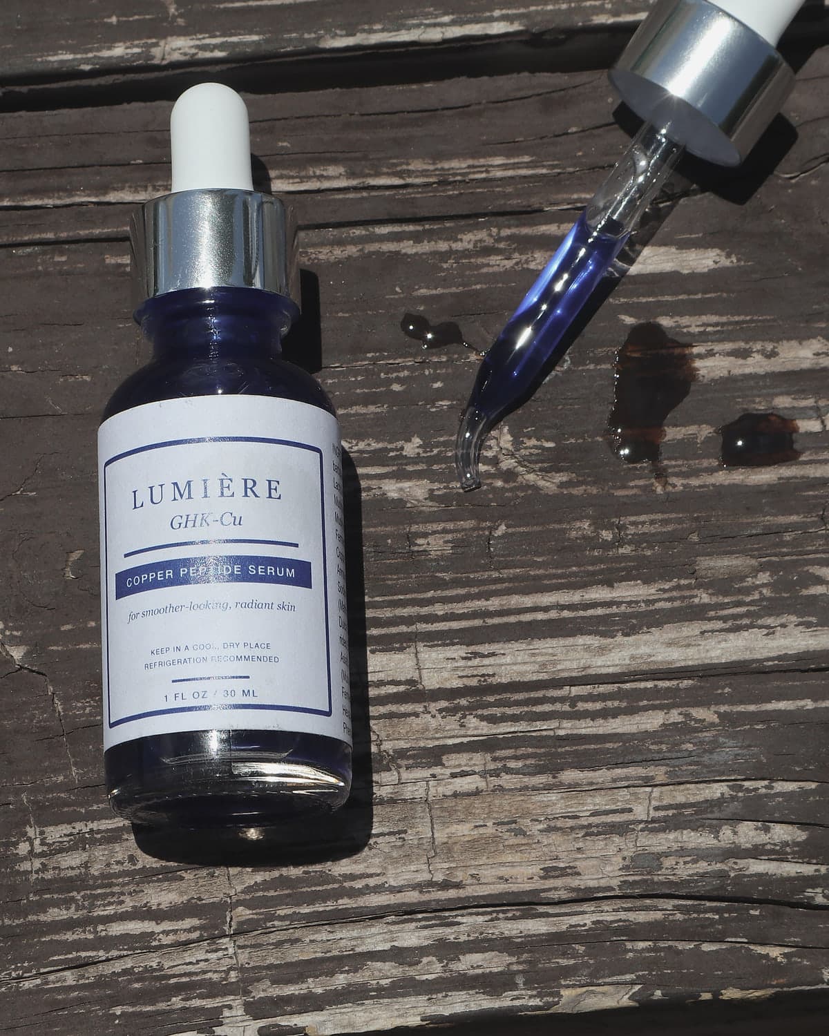 Lumiere Routine GHK-Cu Copper Peptide Serum on weathered wood beside its glass dropper, the deep blue serum visible in the pipette